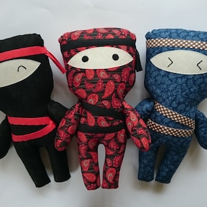 Puede incluir: Tres juguetes ninja de peluche hechos a mano. El ninja de la izquierda es negro con detalles rojos. El ninja del centro es rojo con patrones paisley negros. El ninja de la derecha es azul con detalles marrones.