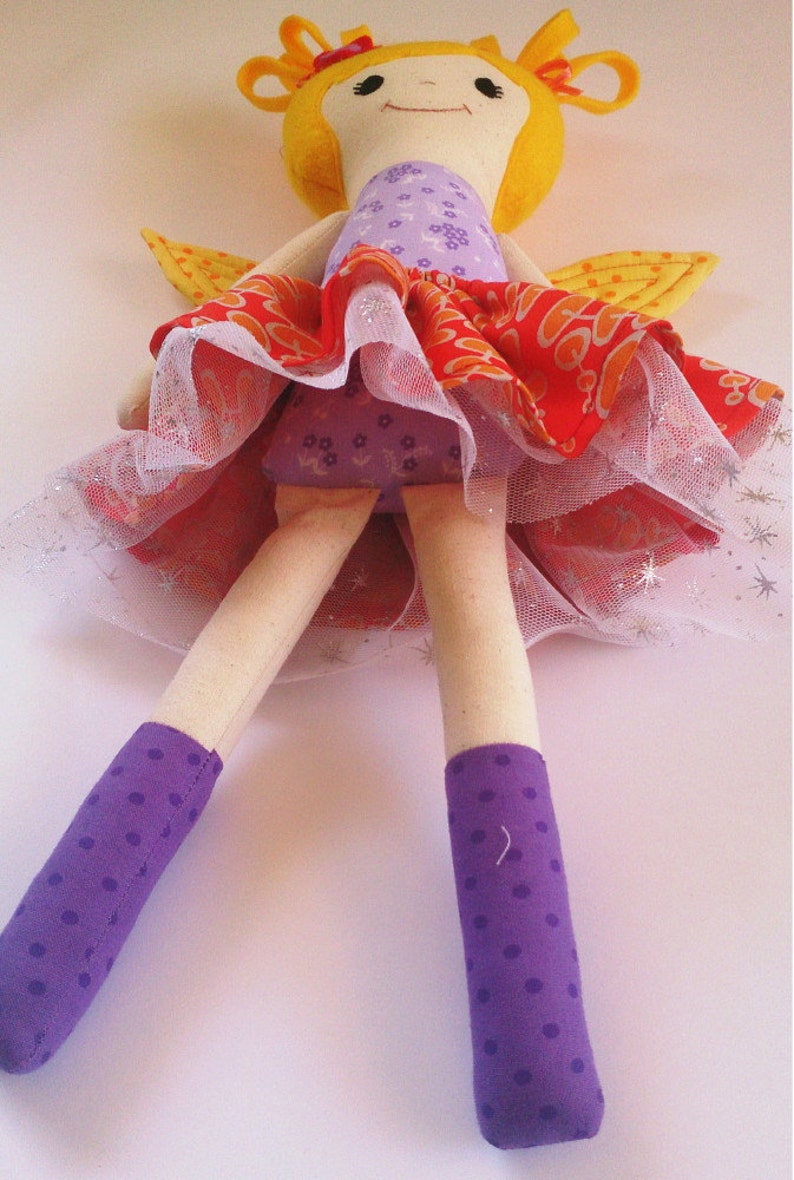 May include: A handmade doll with yellow hair, a purple and orange dress, and purple polka dot socks. The doll is wearing a tulle skirt and has wings.