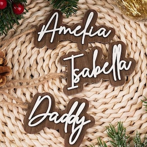 Puede incluir: Etiquetas de nombre de madera con letras blancas en cursiva que deletrean "Amelia", "Isabella" y "Daddy". Las etiquetas son marrones y se muestran sobre un fondo tejido con decoraciones navideñas.