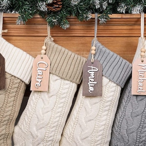 May include: Four cable-knit Christmas stockings in cream, beige, and grey hang from a wooden mantel. Each stocking has a wooden tag with a name: Joshua, Claire, Amelia, and Michael. The tags are suspended by twine and wooden beads.