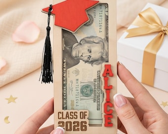 May include: A wooden graduation money holder with a red mortarboard and tassel. The holder contains a twenty-dollar bill and the name "ALICE" in red letters. The text "CLASS OF 2026" is also visible. A white gift box with a gold ribbon is in the background.