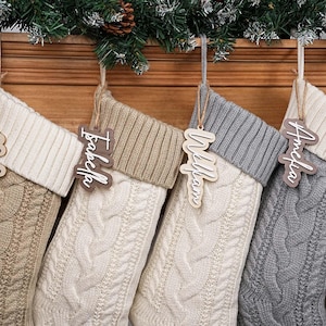 Puede incluir: Cuatro calcetines navideños de punto trenzado en tonos neutros, cada uno con una etiqueta de nombre de madera: Madison, Isabella, William y Amelia. Los calcetines cuelgan de una repisa de madera decorada con ramas de hoja perenne y una piña.