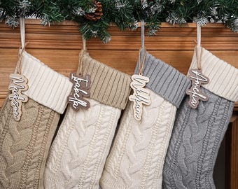 Op de afbeelding: Vier kabelgebreide kerstsokken in neutrale tinten, elk met een houten naamlabel: Madison, Isabella, William en Amelia. De sokken hangen aan een houten schoorsteenmantel versierd met groenblijvende takken en een dennenappel.