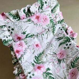 Könnte beinhalten: Ein weißes Babybettwäsche-Set mit einem floralen Muster aus rosa Rosen und grünen Blättern. Die Bettwäsche hat einen gerüschten Rand und ist auf einem weißen Stubenwagenrahmen platziert. Das Blumenmuster wiederholt sich auf Kissen und Decke.