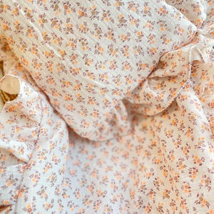 Op de afbeelding: Een witte deken met een bloemenpatroon en een gefranjerde rand. De deken heeft een delicaat ontwerp van kleine oranje en bruine bloemen. De stof lijkt zacht en licht, geschikt voor een baby of kind.