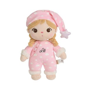 Pode incluir: Boneca de pelúcia em um macacão rosa com padrões de nuvens brancas e touca de dormir combinando. A boneca tem tranças loiras, olhos castanhos grandes e o nome "Lili" impresso no peito. A touca de dormir tem um detalhe em forma de estrela.