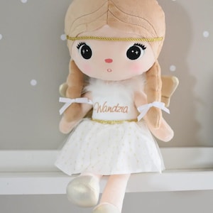 Può includere: Bambola di peluche a forma di angelo, con incarnato beige, capelli biondi e grandi occhi neri. La bambola indossa un vestito bianco con dettagli dorati e il nome "Wandzia". Ha ali dorate e una fascia dorata.