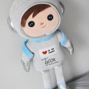 Könnte beinhalten: Eine Plüsch-Astronautenpuppe in Grau-, Silber- und Blautönen. Die Puppe hat ein lächelndes Gesicht mit braunen Haaren und einem silbernen Helm. Auf der Brust der Puppe befinden sich ein Herz und ein Gesichtsdesign mit dem Namen "Tyson" und dem Datum "10.03.2025" darunter.