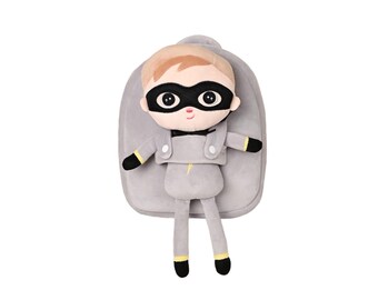 May include: A gray plush backpack with a superhero doll attached. The doll has a black mask, blonde hair, and a gray outfit with yellow accents. The backpack is a solid gray color.