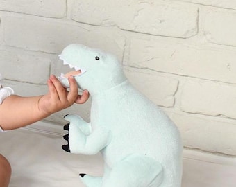May include: A soft, light green plush dinosaur toy with black claws and teeth. The toy is positioned on a white surface against a white brick wall. A small hand is reaching towards the dinosaur's mouth.