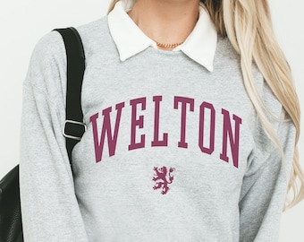 May include: A gray sweatshirt with the word "WELTON" printed in maroon letters. A maroon lion crest is below the text. The sweatshirt is worn by a person with a white collared shirt underneath and a black belt with a double ring buckle.