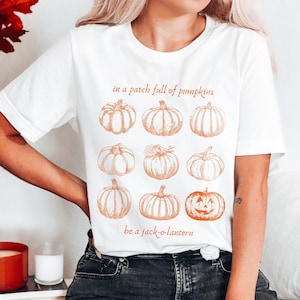 May include: White t-shirt with a pumpkin design. The shirt displays nine sketched pumpkins in orange hues, accompanied by the text "in a patch full of pumpkins be a jack-o-lantern".