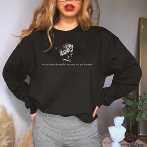 May include: Black crew neck sweatshirt with a white graphic of a classical bust and the text "we are more beautiful because we are doomed". The sweatshirt is a solid black color.