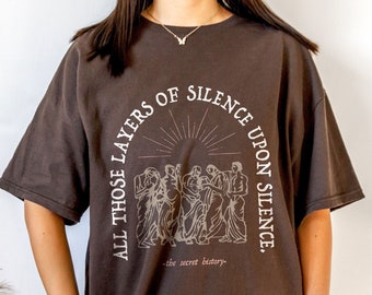 Puede incluir: Una camiseta marrón de gran tamaño con un estampado gráfico de un grupo de personas de pie en círculo con el texto "All those layers of silence upon silence - the secret history -" impreso en la parte delantera.