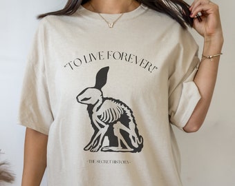 Puede incluir: Una camiseta de gran tamaño de color beige con un gráfico negro de un esqueleto de conejo. El texto "TO LIVE FOREVER!" está por encima del conejo y "THE SECRET HISTORY" está por debajo del conejo.