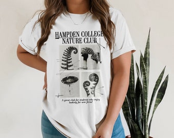 Puede incluir: Camiseta blanca con un estampado gráfico en blanco y negro de helechos y el texto "HAMPDEN COLLEGE NATURE CLUB". El texto debajo del gráfico dice "Un gran club para estudiantes que disfrutan buscando nuevos helechos!"