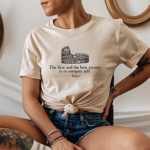 May include: A beige t-shirt featuring a black outline of the Colosseum and the quote "The first and the best victory is to conquer self. -Plato-"