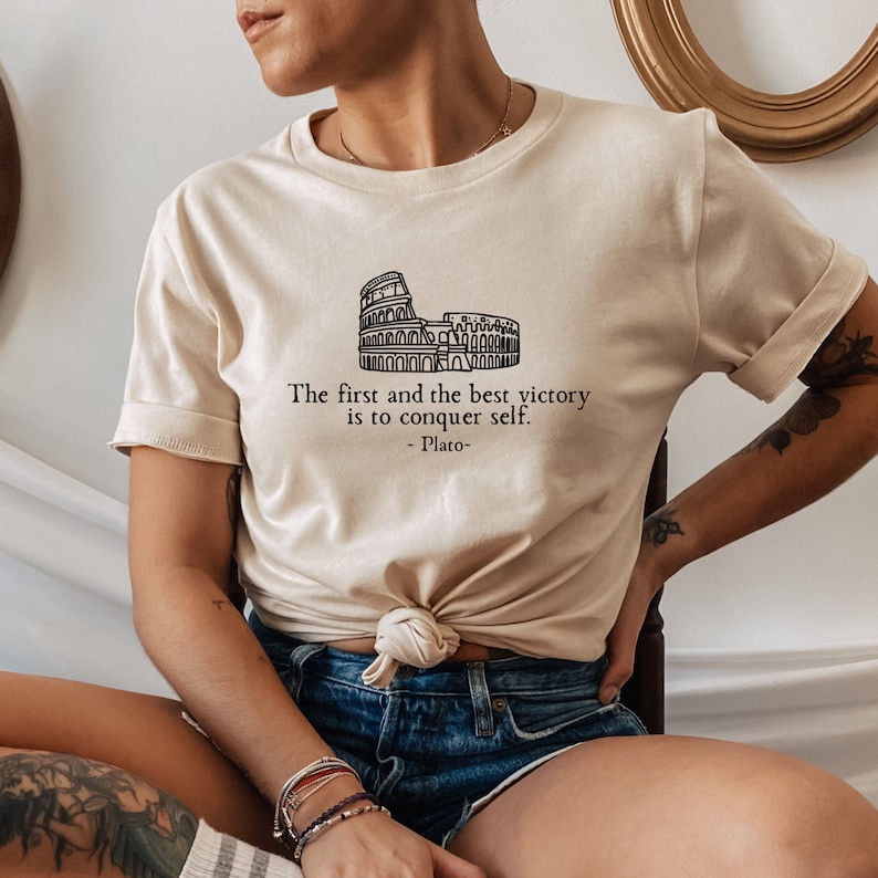 May include: A beige t-shirt featuring a black outline of the Colosseum and the quote "The first and the best victory is to conquer self. -Plato-"