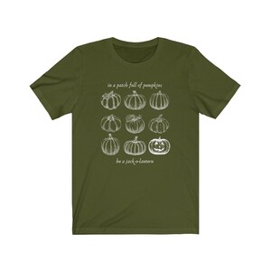 May include: An olive green t-shirt with a white illustration of pumpkins and the text "in a patch full of pumpkins, be a jack-o-lantern". The design features nine pumpkin drawings, including a jack-o-lantern.