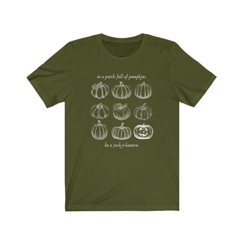 May include: An olive green t-shirt with a white illustration of pumpkins and the text "in a patch full of pumpkins, be a jack-o-lantern". The design features nine pumpkin drawings, including a jack-o-lantern.