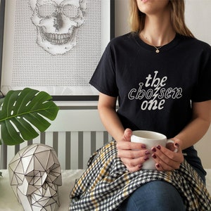 May include: A person wearing a black t-shirt with the text "the chosen one" in white lettering. The person is holding a white mug and sitting on a checkered blanket.