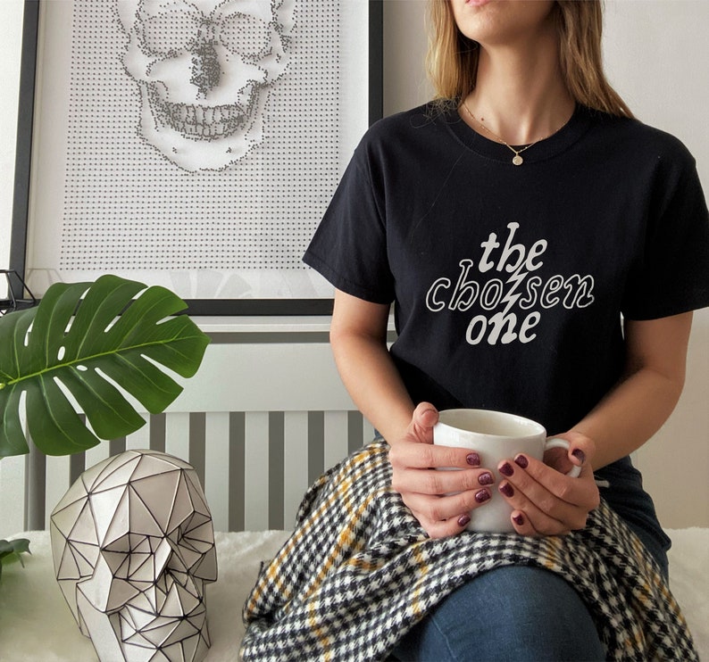 May include: A person wearing a black t-shirt with the text "the chosen one" in white lettering. The person is holding a white mug and sitting on a checkered blanket.