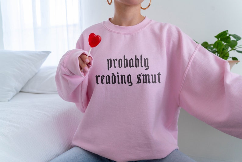 Puede incluir: Sudadera rosa claro con la frase "probably reading smut" en fuente g&oacute;tica negra. Se sostiene un caramelo rojo en forma de coraz&oacute;n. La sudadera es de estilo casual.