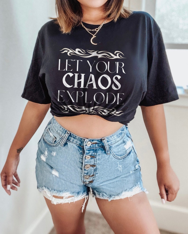 May include: Black t-shirt with the text "LET YOUR CHAOS EXPLODE" in silver lettering. The shirt is paired with light blue denim shorts with a button fly and distressed detailing. A gold necklace with a crescent moon pendant is also visible.