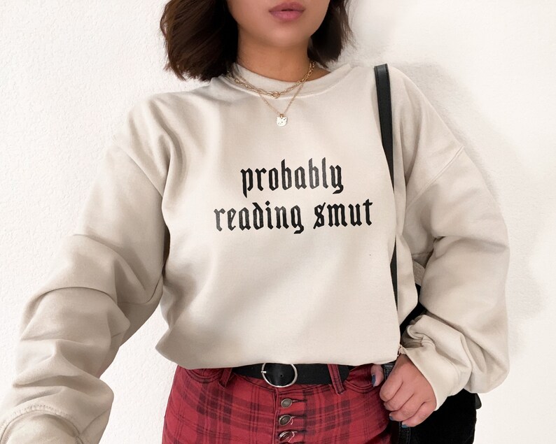 Puede incluir: Sudadera beige con la frase "probably reading smut" impresa en negro con fuente g&oacute;tica. La sudadera tiene mangas largas y cuello redondo. La persona lleva un cintur&oacute;n negro y una falda a cuadros rojos.