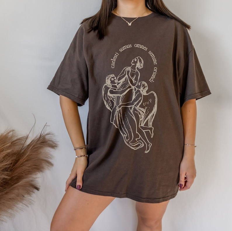Może przedstawiać: Brązowy, oversize'owy t-shirt z nadrukiem graficznym przedstawiającym trzech anioł&oacute;w. Tekst na koszulce brzmi "caelesti sumus omnes semine originis".