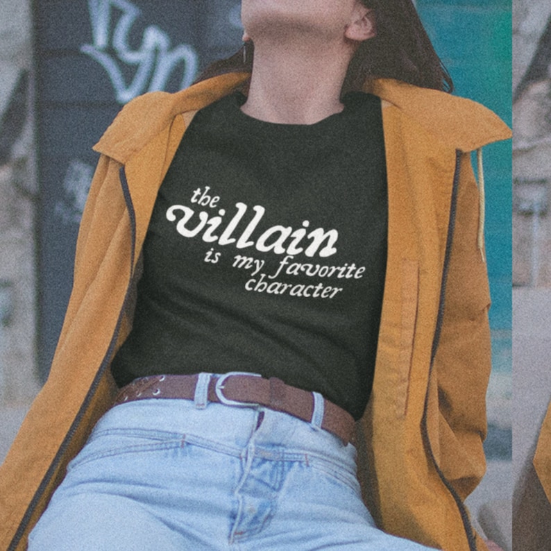 May include: A black t-shirt with white text that reads "the villain is my favorite character". The person wearing the shirt is also wearing a brown jacket and blue jeans.