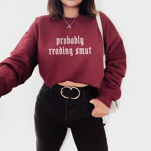 Puede incluir: Una sudadera granate con el texto "probably reading smut" en blanco con fuente g&oacute;tica. La sudadera es de manga larga y corta. La persona lleva pantalones negros y un cintur&oacute;n negro con hebilla dorada.