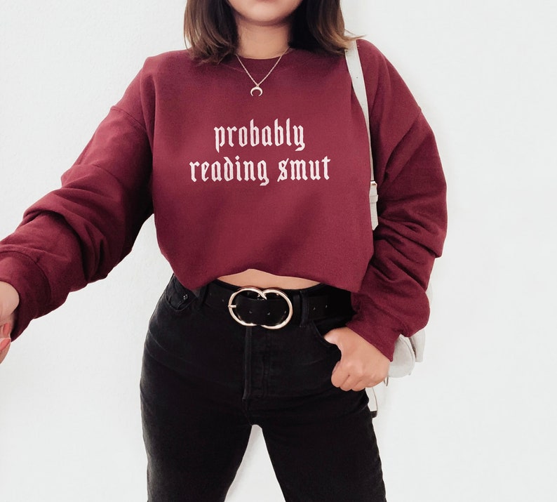 Puede incluir: Una sudadera granate con el texto "probably reading smut" en blanco con fuente g&oacute;tica. La sudadera es de manga larga y corta. La persona lleva pantalones negros y un cintur&oacute;n negro con hebilla dorada.