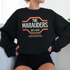 May include: Black sweatshirt with a retro-style graphic design featuring the text "THE MARAUDERS 1971-1978" in white with orange and yellow stripes. The text "PURVEYORS OF AIDS TO MAGICAL MISCHIEF MAKERS" is printed below the graphic.