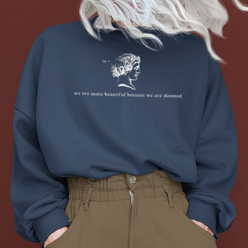 May include: Navy blue sweatshirt with a white graphic of a classical bust and the text "we are more beautiful because we are doomed". The sweatshirt is a relaxed fit and has long sleeves.