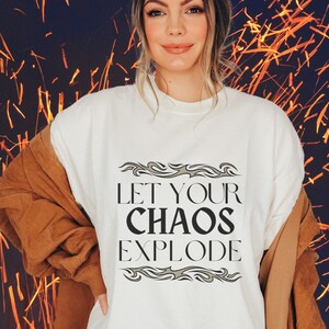 May include: A cream-coloured t-shirt with the text "LET YOUR CHAOS EXPLODE" in black, framed by decorative grey elements. The person is wearing a brown jacket over the shirt.