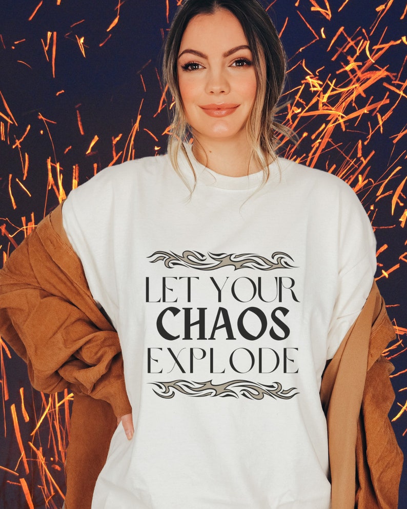 May include: A cream-coloured t-shirt with the text "LET YOUR CHAOS EXPLODE" in black, framed by decorative grey elements. The person is wearing a brown jacket over the shirt.