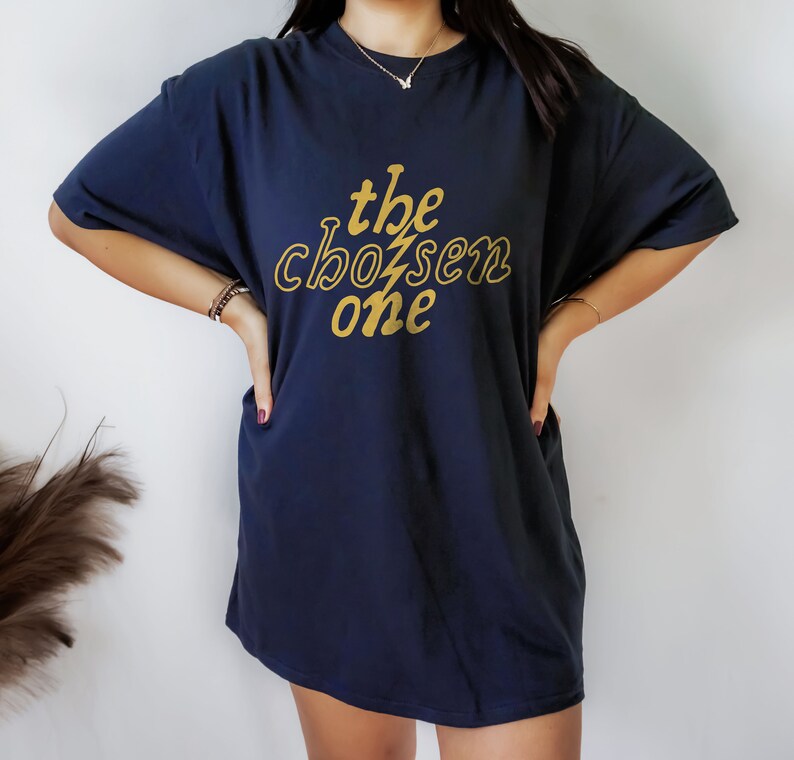 May include: A navy blue oversized t-shirt with a gold graphic that says "the chosen one" in a retro font.
