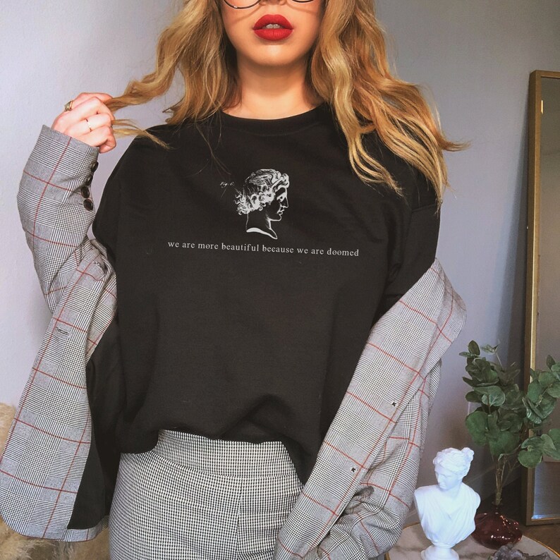 May include: Black crew neck sweatshirt with a white graphic of a classical bust and the text "we are more beautiful because we are doomed". The sweatshirt is paired with a plaid blazer and patterned pants.