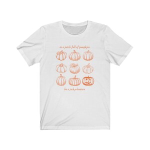 May include: White t-shirt with an orange pumpkin graphic. The design includes nine pumpkins and the text "in a patch full of pumpkins" and "be a jack-o-lantern". The illustration has a vintage style.