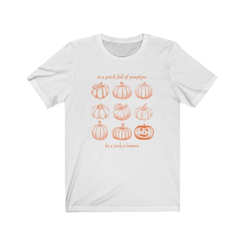 May include: White t-shirt with an orange pumpkin graphic. The design includes nine pumpkins and the text "in a patch full of pumpkins" and "be a jack-o-lantern". The illustration has a vintage style.