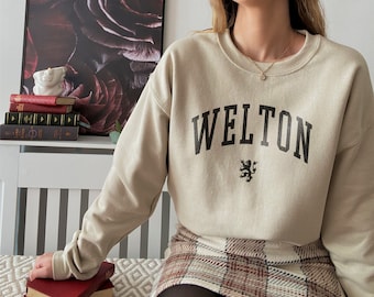 Pode incluir: Camisola bege com a palavra "WELTON" em preto e um pequeno emblema preto. A pessoa está usando uma saia xadrez com um padrão vermelho e marrom.