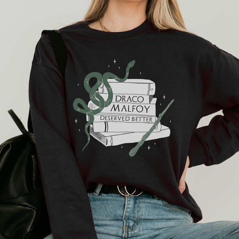 Puede incluir: Sudadera negra con una serpiente verde, una pila de libros y una varita. El texto en los libros dice "Draco Malfoy Deserved Better".