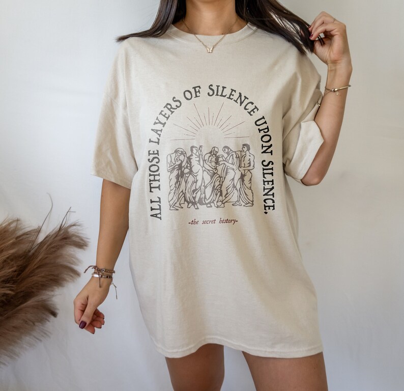May include: A beige oversized t-shirt with a graphic print of a group of people standing in a circle with the text "All those layers of silence upon silence." and "the secret history" underneath.