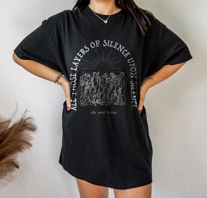 May include: Black oversized t-shirt with a graphic print of a group of people standing in front of a sunburst. The text reads "All those layers of silence upon silence - the secret history -"