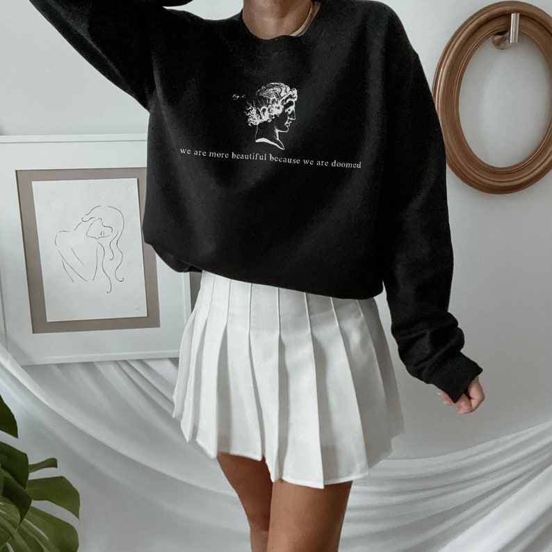 May include: Black sweatshirt with a white graphic of a classical bust and the text "we are more beautiful because we are doomed." Paired with a white pleated tennis skirt. The sweatshirt has long sleeves.