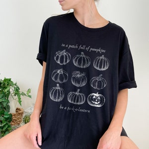 May include: Black t-shirt with a white illustration of pumpkins, including a jack-o'-lantern. The text reads "in a patch full of pumpkins" and "be a jack-o'-lantern". A casual, comfortable top for autumn.