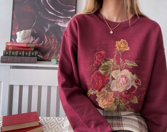Może przedstawiać: Bordowa bluza z nadrukiem kwiatowym przedstawiającym róże, liście i odręczne pismo. Bluza ma okrągły dekolt i długie rękawy.