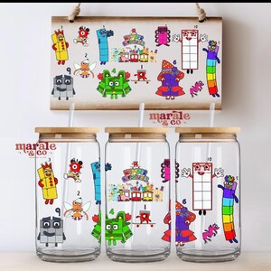 Può includere: Tre bicchieri di vetro trasparente con coperchi in legno e cannucce, decorati con personaggi numerici di cartoni animati colorati. Un cartello di legno sopra mostra personaggi simili. I bicchieri hanno il testo "marale & co."