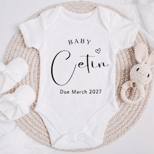 May include: A white short-sleeved baby bodysuit featuring the black printed text BABY Cetin with a small heart icon and the text Due March 2027. The bodysuit is accompanied by a pair of white baby booties and a small crocheted rabbit rattle toy.